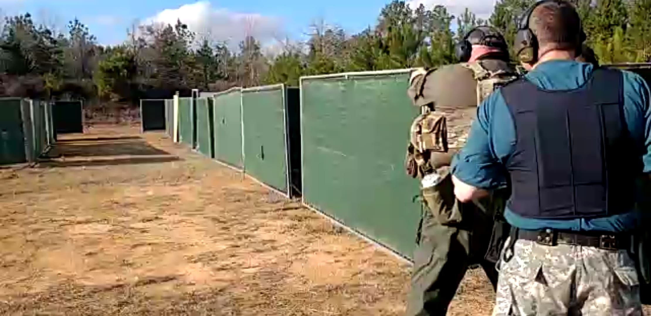 Two people in tactical gear and headphones are at an outdoor shooting range with high green barriers.