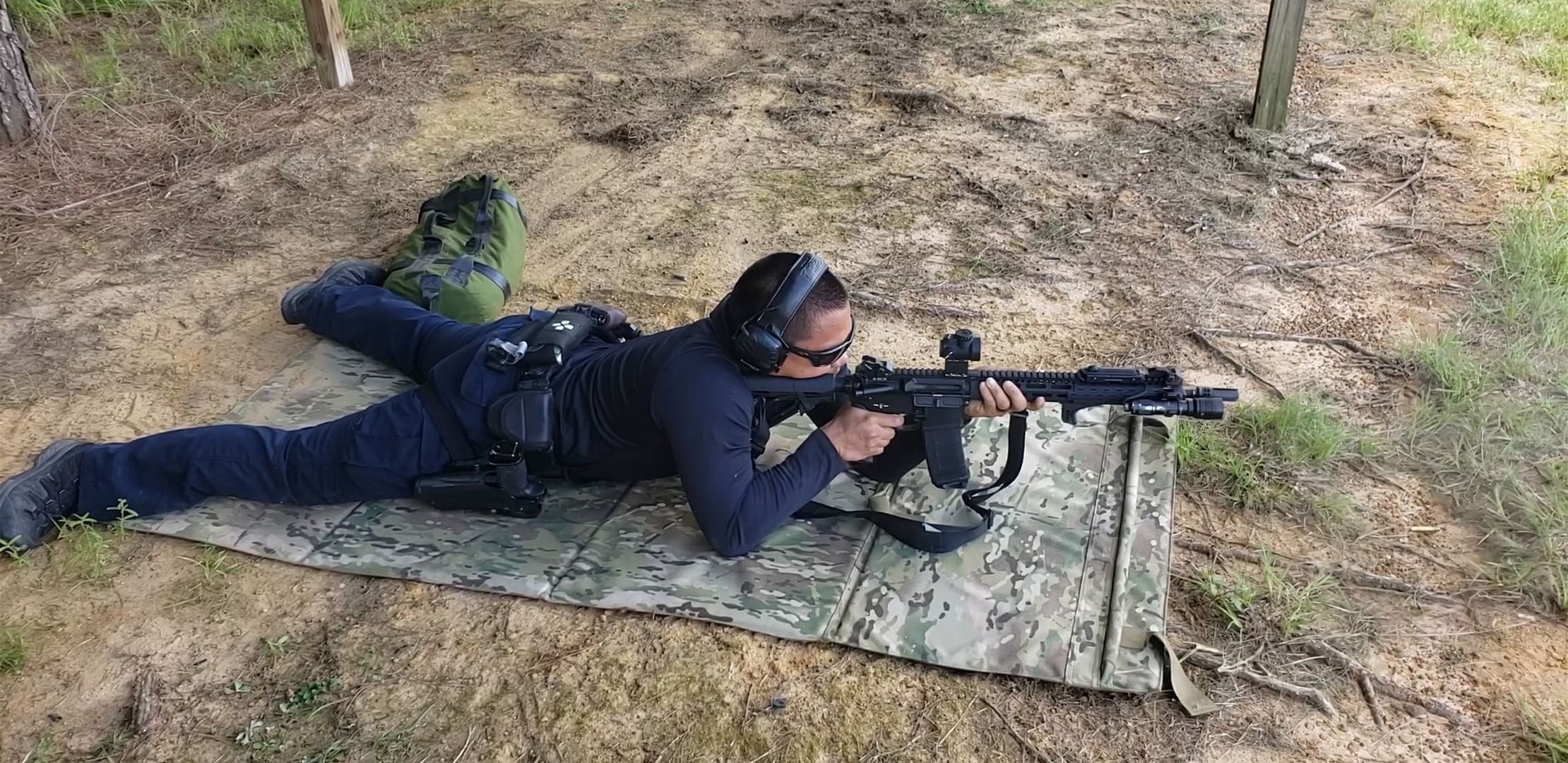 A person lying prone on a camouflage mat, focusing intently while aiming a rifle with a scope.